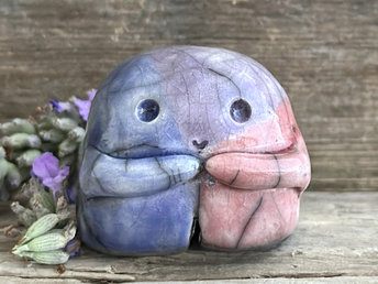 A sweet little nature spirit ceramic guardian statue glazed in the bisexual flag pink, purple, and blue colours and with a lovely, gentle face and little arms. It has a small branch design carved up one side.