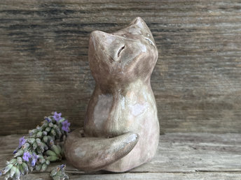 a beautiful Shinto Inari kitsune fox kami spirit ceramic raku statue. it's looking up with a peaceful, happy expression on its gentle face and is glazed in soft browns and pink-browns with its tummy being lighter than the rest. its tail is curled around its body on its right side, and it has white, snow-like marks in the glaze on the left side of its head and back.