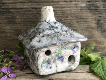 Raku ceramic kurinuki cottage glazed in white with green, purple, and violet flower patterns on the walls. The roof has a chimney and is glazed in grey, white, and black to look like slate. It has a cute little doorway and lots of windows.