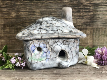 Raku ceramic kurinuki cottage glazed in grey with green and blue-violet flower patterns on the walls. The roof has a chimney and is glazed in grey to look like stone tiles. It has a cute little doorway and lots of windows.