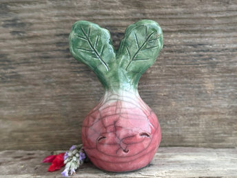 a very smiley and happy radish spirit raku ceramic sculpture. it has a round body glazed in a soft radish red colour. the top of its body fades to white, and it has two green leaves on the top of its head. body. it is round with green leaves on top!