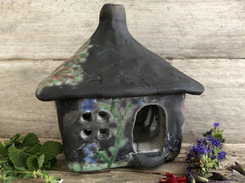 Raku ceramic kurinuki cottage glazed with a tree and lots of flowers and vines in different colours. The house is left natural, unglazed black apart from where the designs are. The roof has a chimney, and the house has a cute little doorway and lots of windows.
