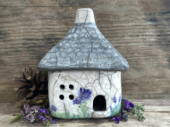 Rustic raku ceramic kurinuki cottage glazed in white with green plant and purple, violet, and indigo flower patterns on the walls. The roof has a chimney and is glazed in grey with hints of white to look like slate. It has a cute little doorway and lots of windows.