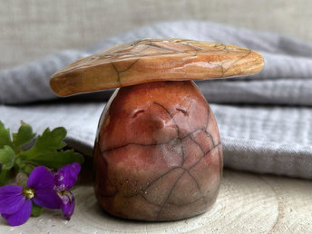 Raku ceramic sculpture glazed in a bottom-to-top gradient of brown to bordeaux red to red-orange with a kind, contented face. It has a golden yellow and orange leaf on its head.