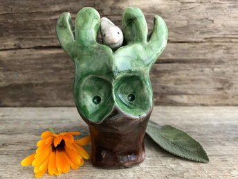 Raku ceramic sculpture of a tree spirit glazed in brown at the bottom and green at the top. He has a face and branches and a little white bird who can sit with him in his branches or on the ground beside him.