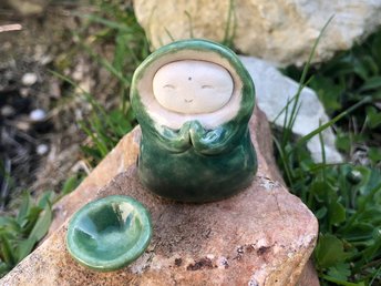 Ceramic sculpture of a little praying forest monk. His sweet face is unglazed white and he wears a green winter robe with a big hood. He has little light green mittens on his hands and a bowl to match.