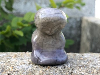 Raku ceramic sculpture of a Jizo or Kuan Yin spirit with its head bowed and its hands in its lap. It is glazed in soft grey and violet and looks very peaceful and meditative.
