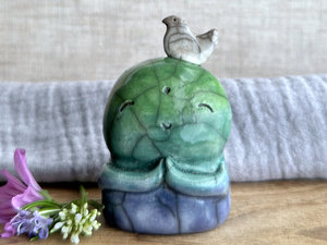 a raku ceramic jizo nature spirit guardian sculpture glazed in a bottom-to-top gradient of blue to forest green to spring green. they have a happy, smiley face, little praying hands, and a sweet white bird on their head.