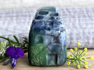 Tiny raku ceramic cottage glazed in shades of blue, green, and teal. Its roof is carved to look like slates. It has a carved door and windows and lots of plant and flower carvings on its walls.