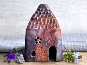 Raku ceramic kurinuki cottage glazed in bordeaux red, warm orange, mauve, and purple. It has a cute little chimney, plant and flower carvings on its walls, a cute door, and little windows, one of which is carved in a flower shape. Its roof is carved to look like tiles.