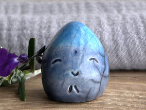 a raku raindrop sculpture with a happy face and two carved little praying hands. it's glazed in a bottom-to-top gradient of blue to turquoise.