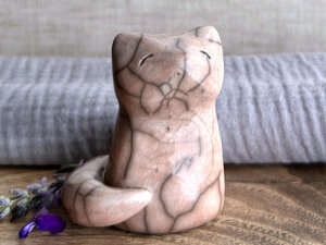A Shinto Inari kitsune fox spirit ceramic raku statue glazed in soft, warm peach colour with lots of raku crackles. Its tail is curled around the right side of its body, and it has a kind face.