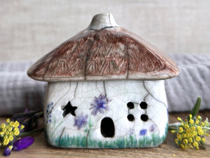 a rustic raku ceramic kurinuki cottage glazed in white with green plant carvings and purple, violet, and indigo flower carvings on the walls. the roof has a chimney and is glazed in brown and carved to look like thatch. the cottage has a cute little doorway and lots of windows including one in a star shape.