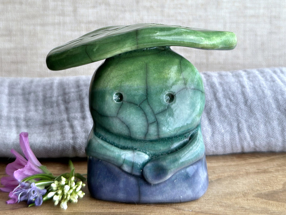 Raku ceramic sculpture glazed in a bottom-to-top gradient of blue to forest green to leaf green with a kind, but somewhat intense and worried, face, and a leaf-green leaf on its head. It also has two cute little arms.