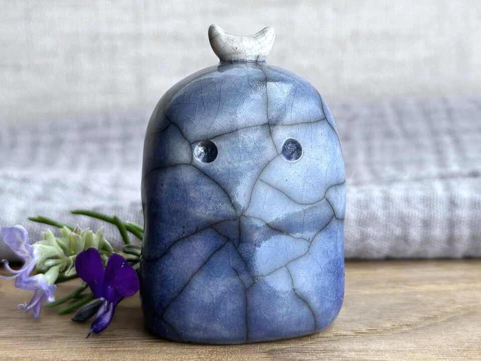 A kodama nature and forest spirit kami ceramic raku sculpture glazed in a bottom-to-top gradient of darker to lighter blue. It has a gentle face, and a white crescent moon on its head.