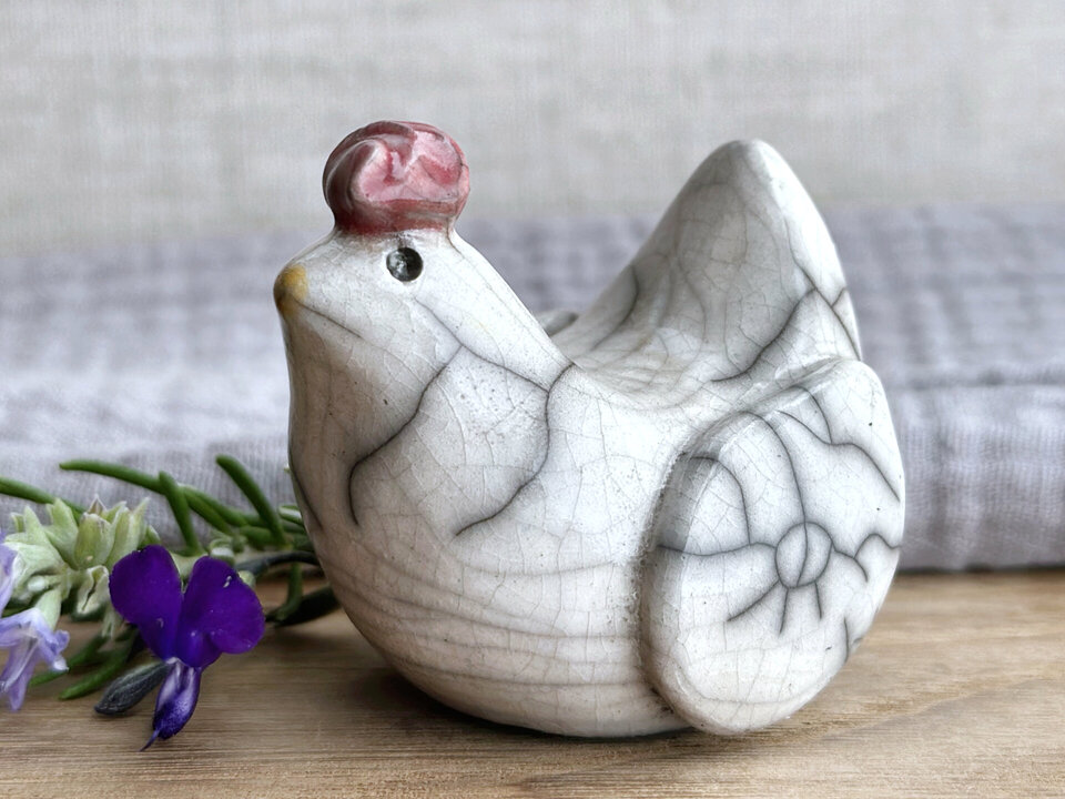 a white raku ceramic chicken with grey eyes, a pale golden-yellow beak, and a bordeaux red comb on the top of her head. she looks very sweet and a little uncertain.