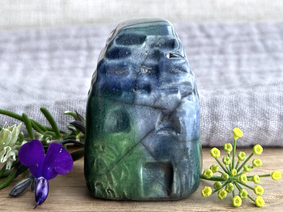Tiny raku ceramic cottage glazed in shades of blue, green, and teal. Its roof is carved to look like slates. It has a carved door and windows and lots of plant and flower carvings on its walls.