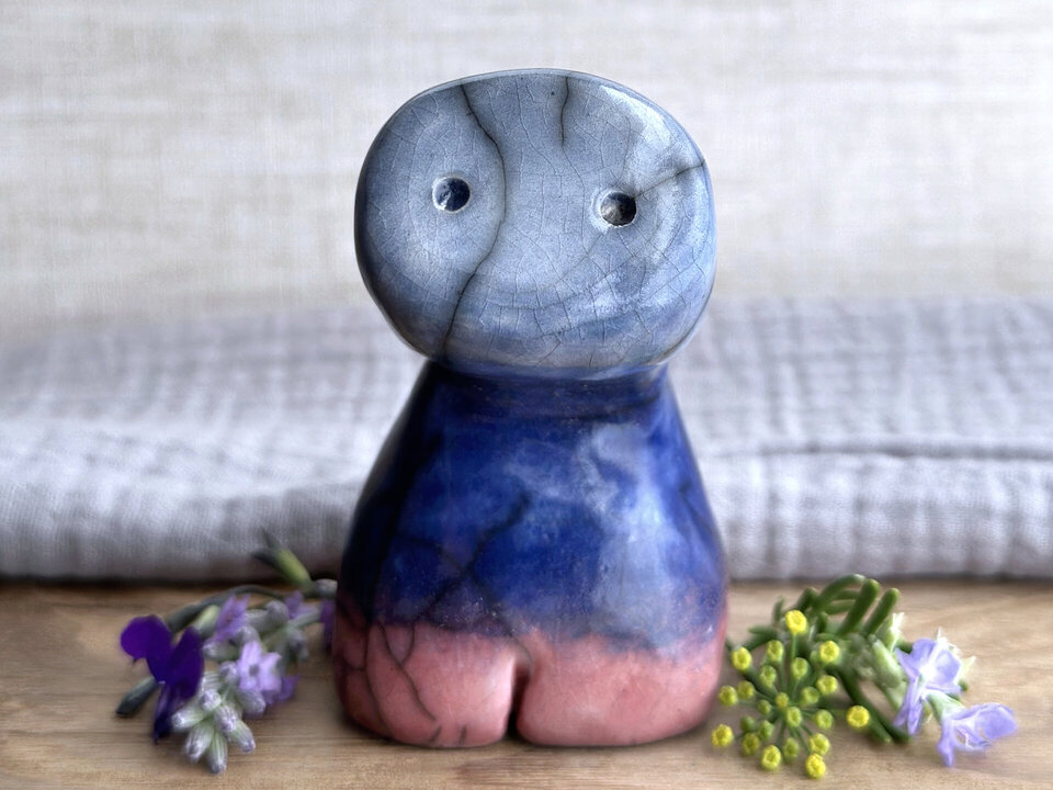 a raku ceramic creature. it has four stubby legs, a widish lower half, a long, tall neck that curves forward, and a roundish head with two round eyes in its flat face. it's glazed in a bottom-to-top gradient of soft pinky red to dark blue to blue. it is a friend.