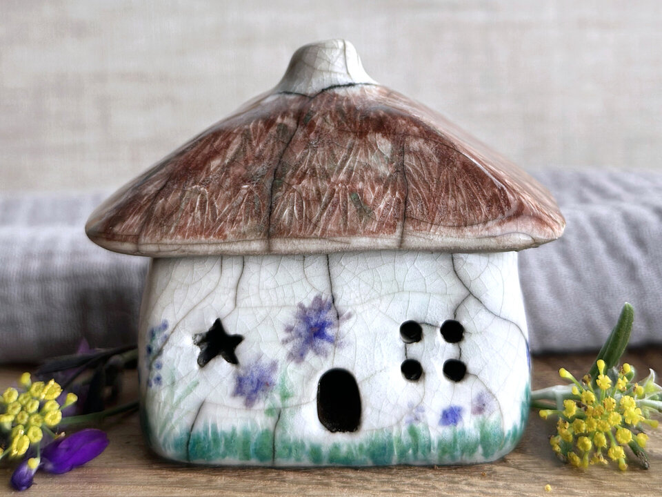 a rustic raku ceramic kurinuki cottage glazed in white with green plant carvings and purple, violet, and indigo flower carvings on the walls. the roof has a chimney and is glazed in brown and carved to look like thatch. the cottage has a cute little doorway and lots of windows including one in a star shape.