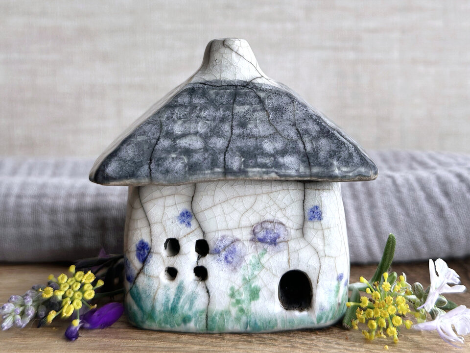 a rustic raku ceramic kurinuki cottage glazed in white with green plant carvings, blue star carvings, and purple, violet, and indigo flower carvings on the walls. the roof has a chimney and is glazed in grey and carved to look like slates. the cottage has a cute little doorway and lots of windows, including one in a crescent moon shape on one of its sides.