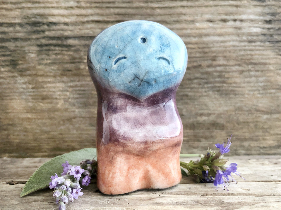 raku ceramic sculpture of an standing elemental sky spirit. it has the impression of arms and legs, and its kind, friend face is flat and shaped like a very wide inverted teardrop. it's glazed in a bottom-to-top gradient of warm burnt-orange to purple to sky blue.