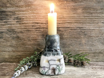 Raku ceramic candlestick in the shape of a house. The top is grey and carved to look like slates. The rest is a white cottage with windows, a door, and flower and plant carvings.