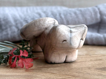 Raku ceramic kitsune fox sculpture glazed in a very pale peach colour. It has a kind, gentle face, and its tail is curled over its back on its right.