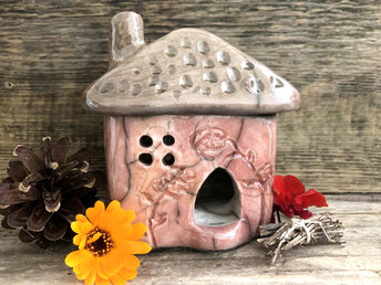 Raku ceramic kurinuki cottage glazed in burnt orange, deep red, and violet with carved flower and tree patterns on the walls. The roof has a chimney, patterning, and is glazed in grey-brown. This house has a cute little doorway and lots of windows.