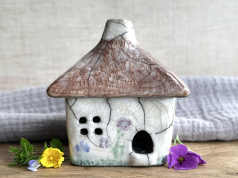a rustic raku ceramic kurinuki cottage glazed in white with green plant carvings and purple, violet, and indigo flower carvings on the walls. the roof has a chimney and is glazed in brown with hints of white and carved to look like thatch. the cottage has a cute little doorway and lots of windows.