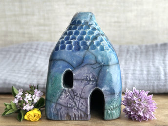 Raku ceramic kurinuki cottage glazed in forest green, blue, indigo, and violet. It has a cute little chimney, plant carvings on its walls, a cute door and lots of little windows, one of which is in the shape of a crescent moon.