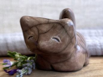 Sitting raku ceramic kitsune fox sculpture glazed in warm brown with hints of mauve. It has a very kind, gentle face, and its tail is curled up its back. It has four stubby legs.