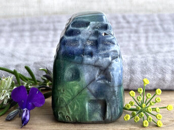 Tiny raku ceramic cottage glazed in shades of blue, green, and teal. Its roof is carved to look like slates. It has a carved door and windows and lots of plant and flower carvings on its walls.