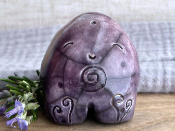 round and vaguely egg-shaped nature spirit sculpture. it is glazed in a top-to-bottom gradient of purple to violet and has spiral carvings on its body. it also has a gentle, happy face.